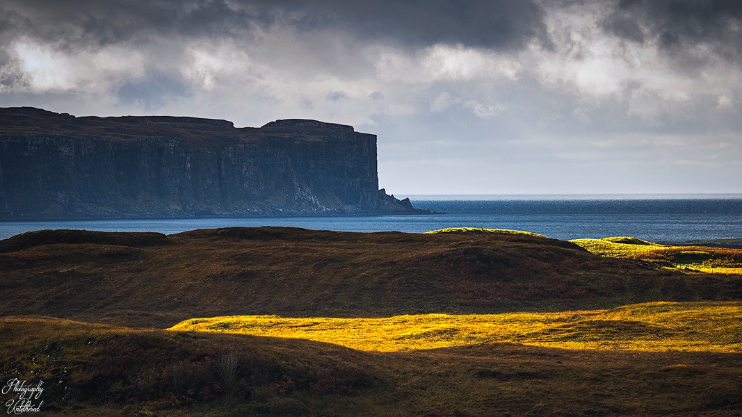 Majestic coastal landscape with dramatic cliffs, dark sea, and cloudy sky. Sunlight highlights golden grass on rolling hills, creating a serene tone. Minginish Peninsula Loch Bracadale Isle of Skye.
