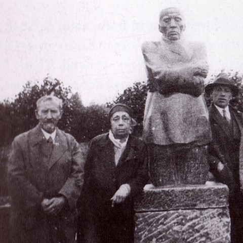 Three men stand beside a stone statue of a man with folded arms. The statue rests on a plinth. The background shows shrubbery, creating a solemn tone. Käthe and Karl Kollwitz Roggeveld German Military Cemetery.