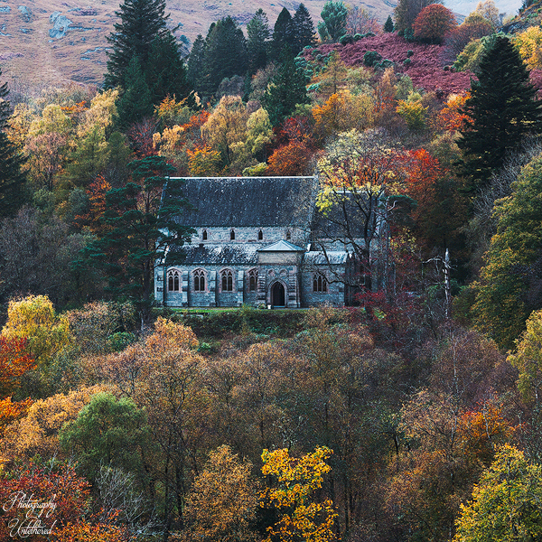 A stone chapel nestled among vibrant autumn trees, with hues of orange, yellow, and green, conveys a serene, picturesque countryside ambience—Church of St Mary and St Finnan, located in Glenfinnan.